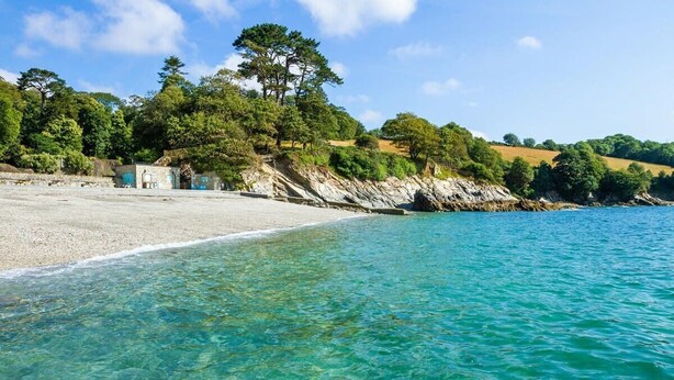 A beach with clear turquoise water, a sandy shore, and a rocky cliff with trees. - Homely Cottage with Sun Terrace, Short Walk to Beach (Porthleven)