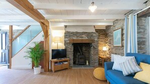 A living room with a stone fireplace, a television on a wooden stand, a blue sofa, and a staircase. - Homely Cottage with Sun Terrace, Short Walk to Beach (Porthleven)