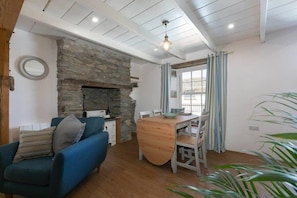 A cozy living room with a stone fireplace, a blue sofa, a wooden dining table, and a large window with curtains. - Homely Cottage with Sun Terrace, Short Walk to Beach (Porthleven)