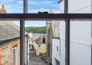 A narrow street with buildings on either side, a clear sky, and a distant view of a harbor with boats. - Sundance Cottage, Central Fowey with Sea Views! (Cornwall)