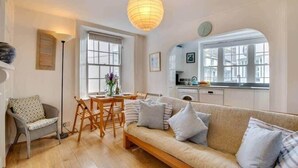 A cozy living room with a sofa, a dining table, and chairs. There is a kitchen area with cabinets and a window. - Sundance Cottage, Central Fowey with Sea Views! (Cornwall)