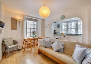 A cozy living room with a sofa, a dining table, and chairs. There is a kitchen area with cabinets and a window. - Sundance Cottage, Central Fowey with Sea Views! (Cornwall)