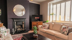 Una sala de estar con chimenea, un televisor mostrando 'NETFLIX', un sofá con cojines estampados y un espejo grande en la pared.