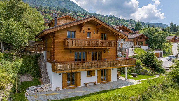 A two-story wooden chalet with a balcony, surrounded by lush greenery and mountains in the background. - Chalet Le Papillon, sauna, 4 Vallées (Haute-Nendaz)