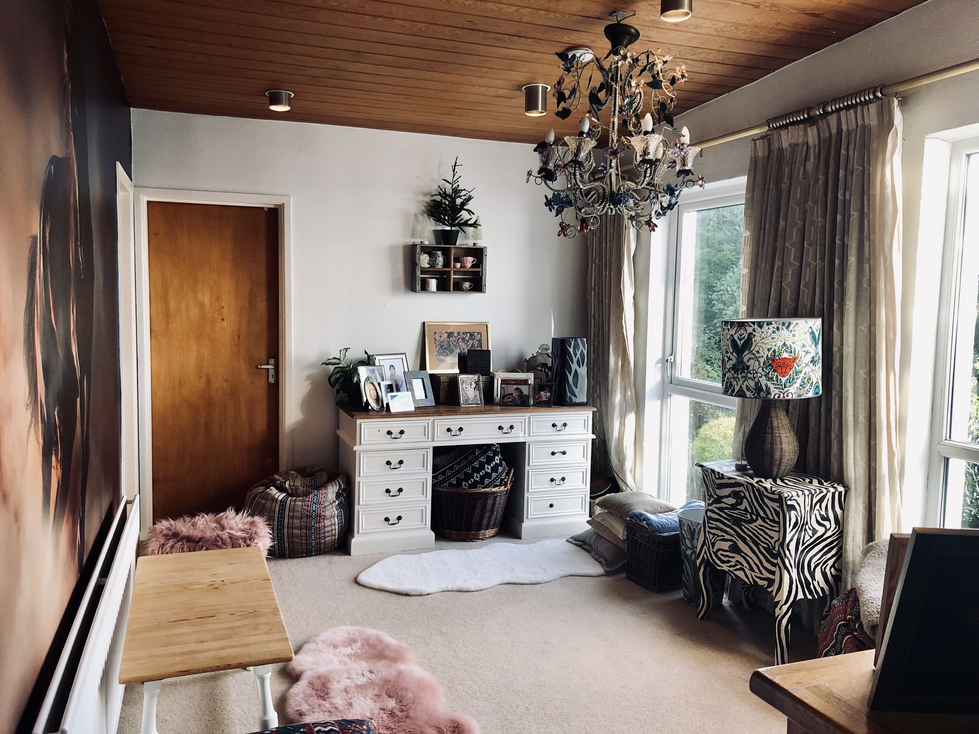 A cozy living room with a wooden ceiling, a chandelier, a white cabinet with drawers, a window with curtains, and a zebra-patterned chair.