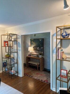 Una habitación con piso de madera, una consola con una lámpara, un cuadro y dos estanterías de vidrio con libros y objetos decorativos.