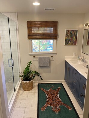 A bathroom with a glass shower enclosure, a window with blinds, a towel rack, a sink with a mirror, and a tiger rug on the floor.