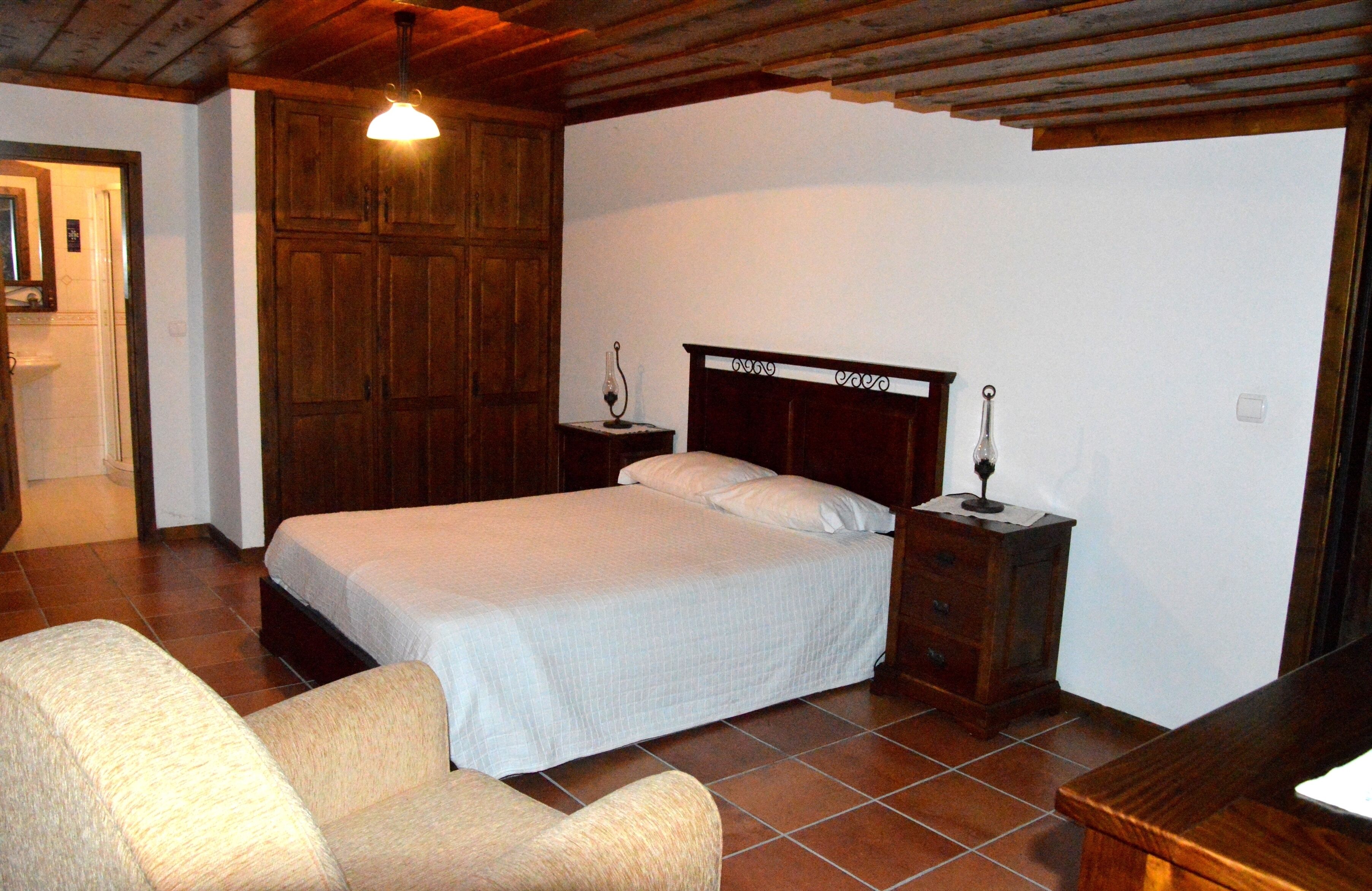 A bedroom with a wooden bed, a nightstand, a chair, and a wooden ceiling.