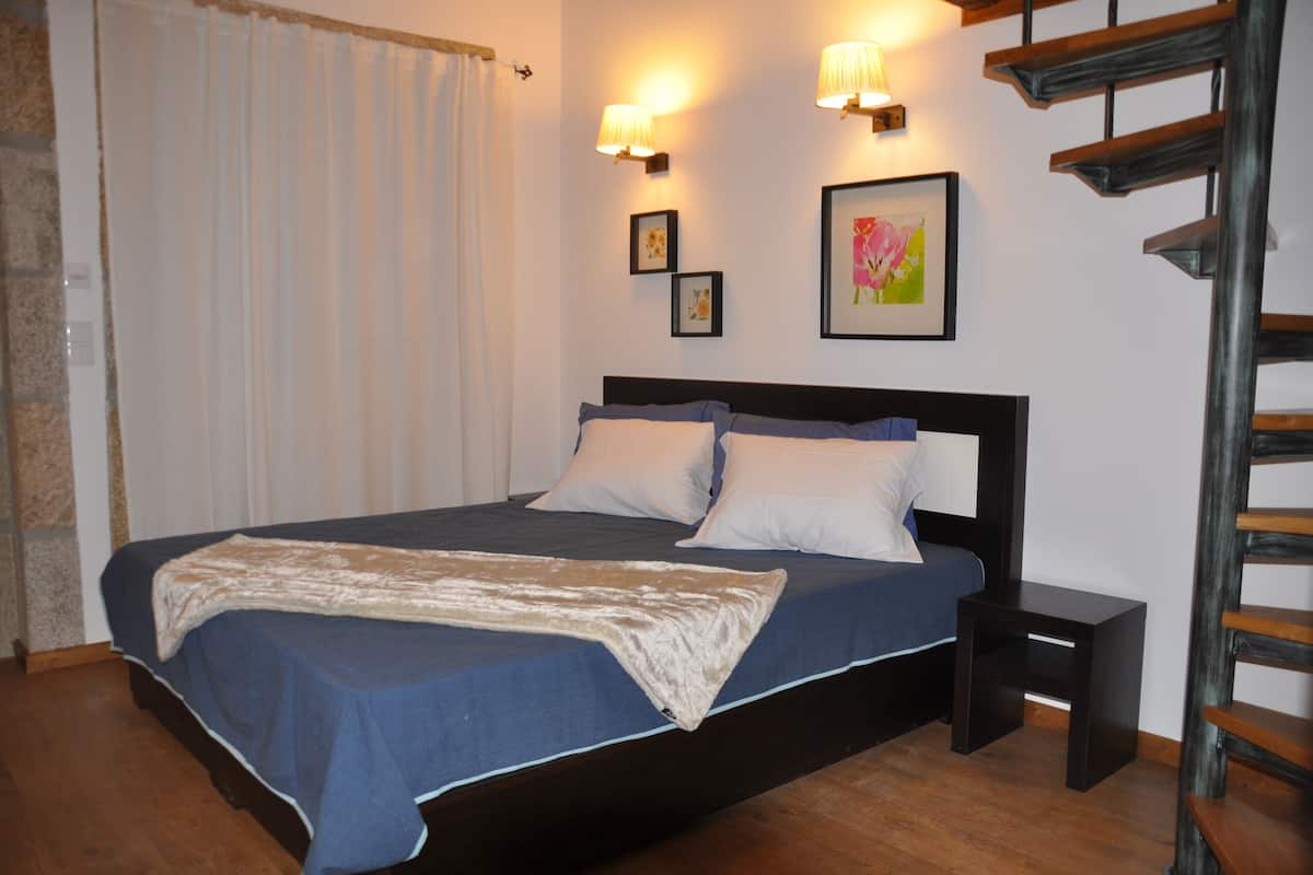 A bedroom with a bed, two bedside tables, a nightstand, a wooden staircase, and a window with curtains.