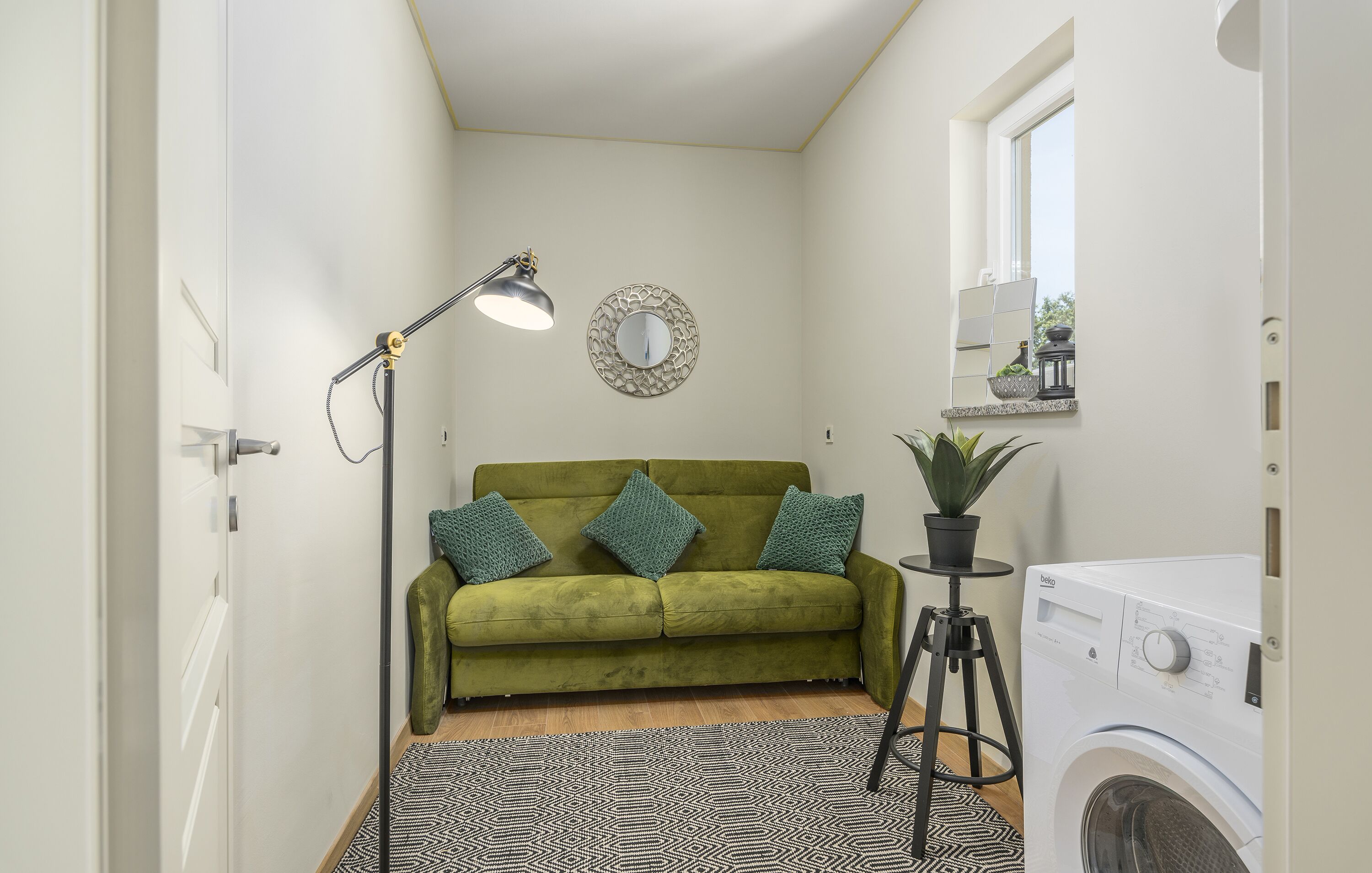 Une petite buanderie comprenant un lave-linge, un canapé vert agrémenté de coussins décoratifs, une lampe sur pied et une petite plante en pot.