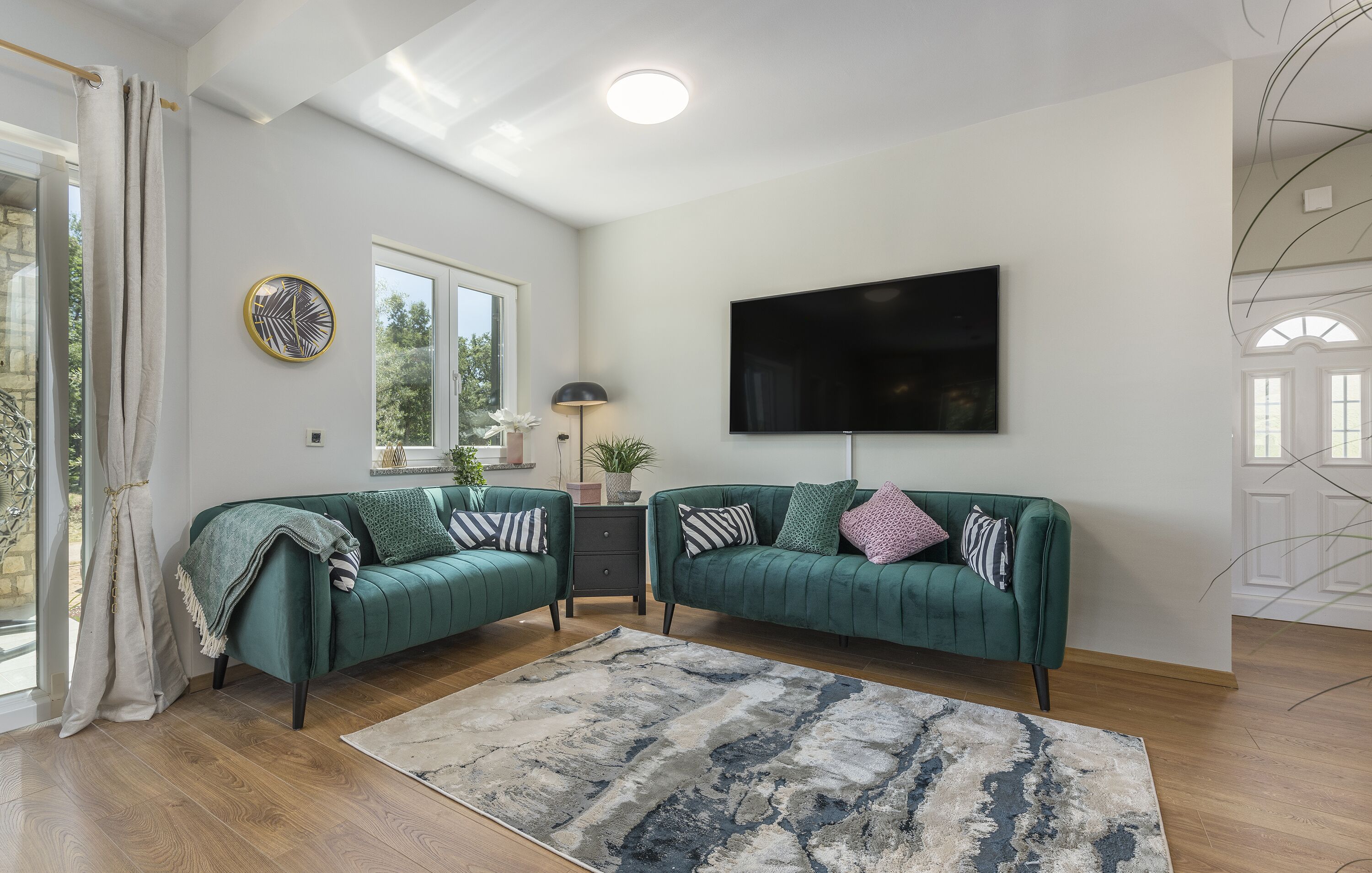Un salon moderne avec deux canapés verts, un grand téléviseur à écran plat, un tapis à motifs et une fenêtre donnant sur un paysage verdoyant.