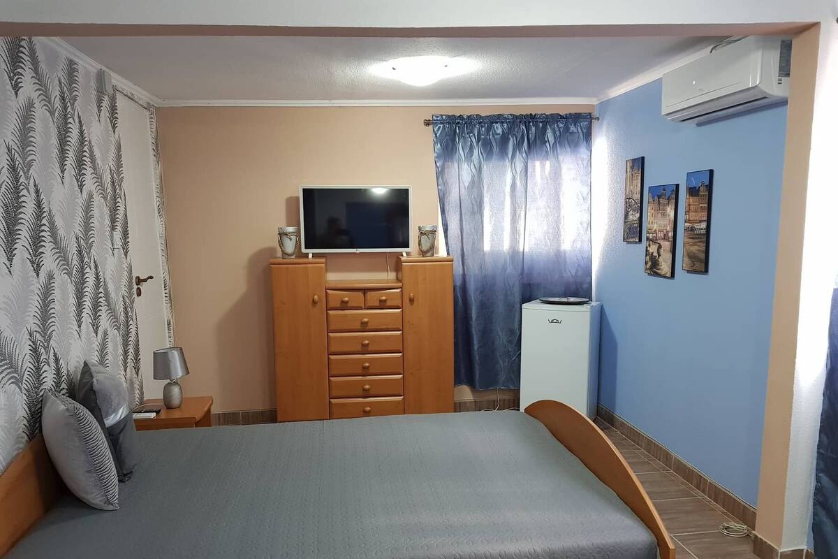 A bedroom with a bed, a TV on a wooden cabinet, a small white appliance, and a window with curtains.