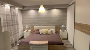 A modern bedroom with a wooden bed, two bedside tables with lamps, a large window with curtains, and a herringbone-patterned floor. - Airport16 B&B (Vecsés)