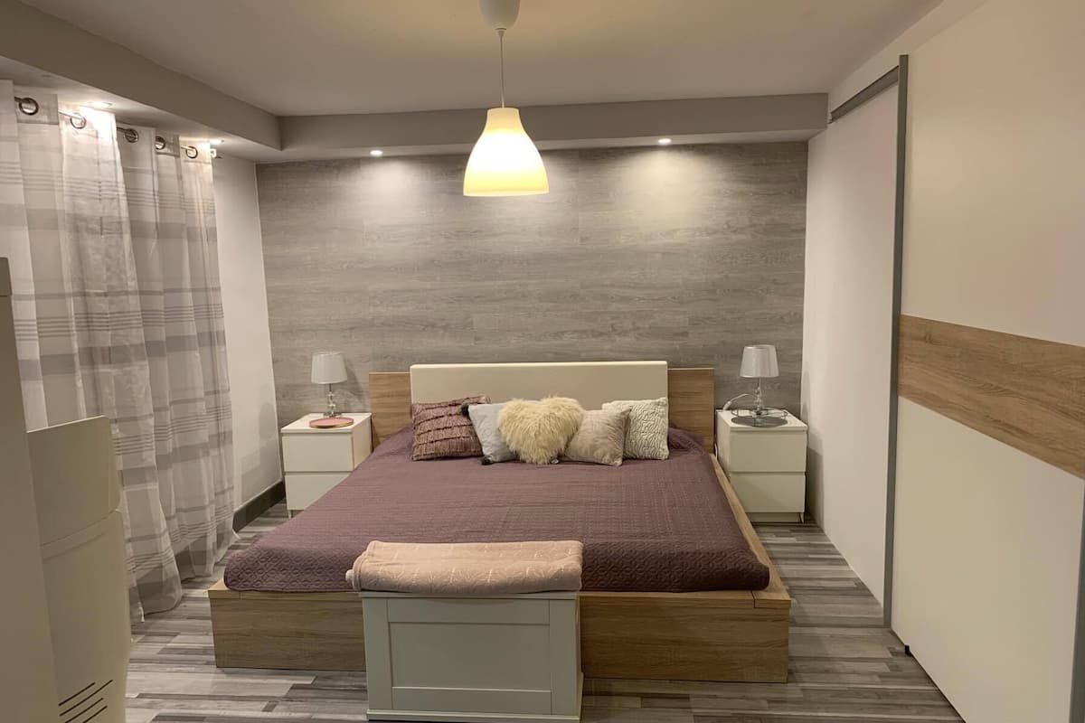A modern bedroom with a wooden bed, two bedside tables with lamps, a large window with curtains, and a herringbone-patterned floor.