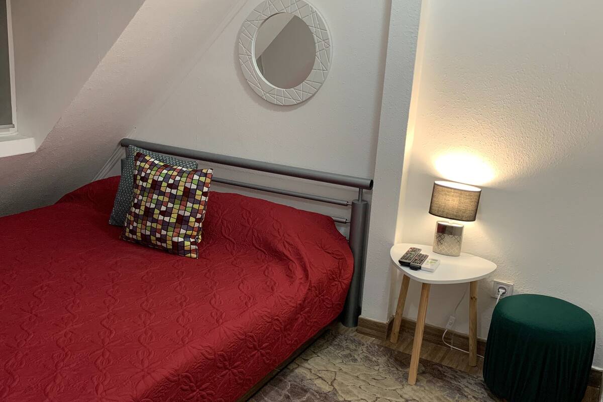 A bedroom with a red bed, a patterned pillow, a small bedside table with a lamp, a green ottoman, and a stone-patterned rug.