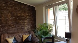 A living room with a stone wall, a brown leather sofa with cushions, a dining table with chairs, and a window with curtains. - Airport16 B&B (Vecsés)