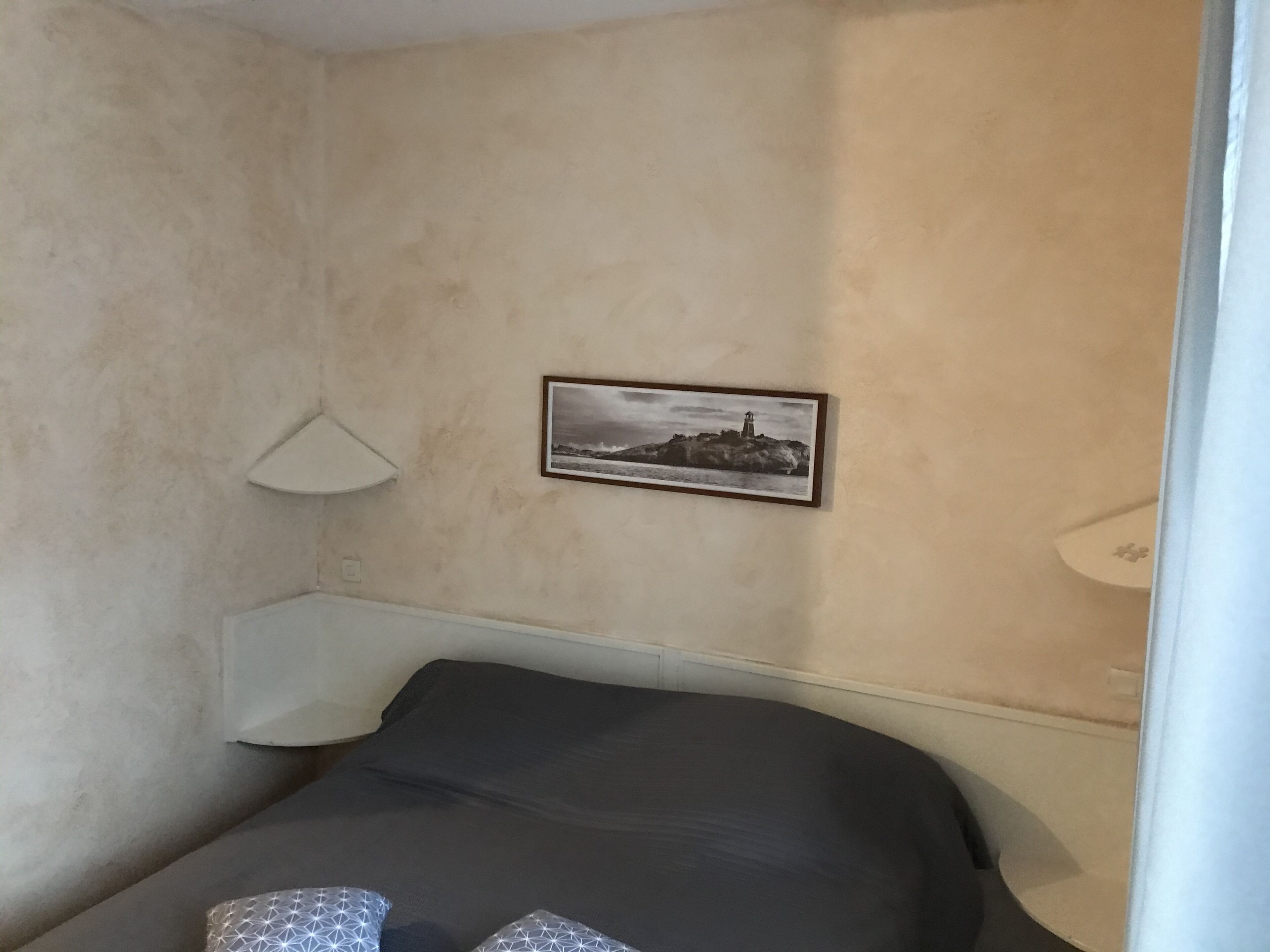 A bedroom with a bed, a wall-mounted lamp, and a framed picture of a coastal scene.