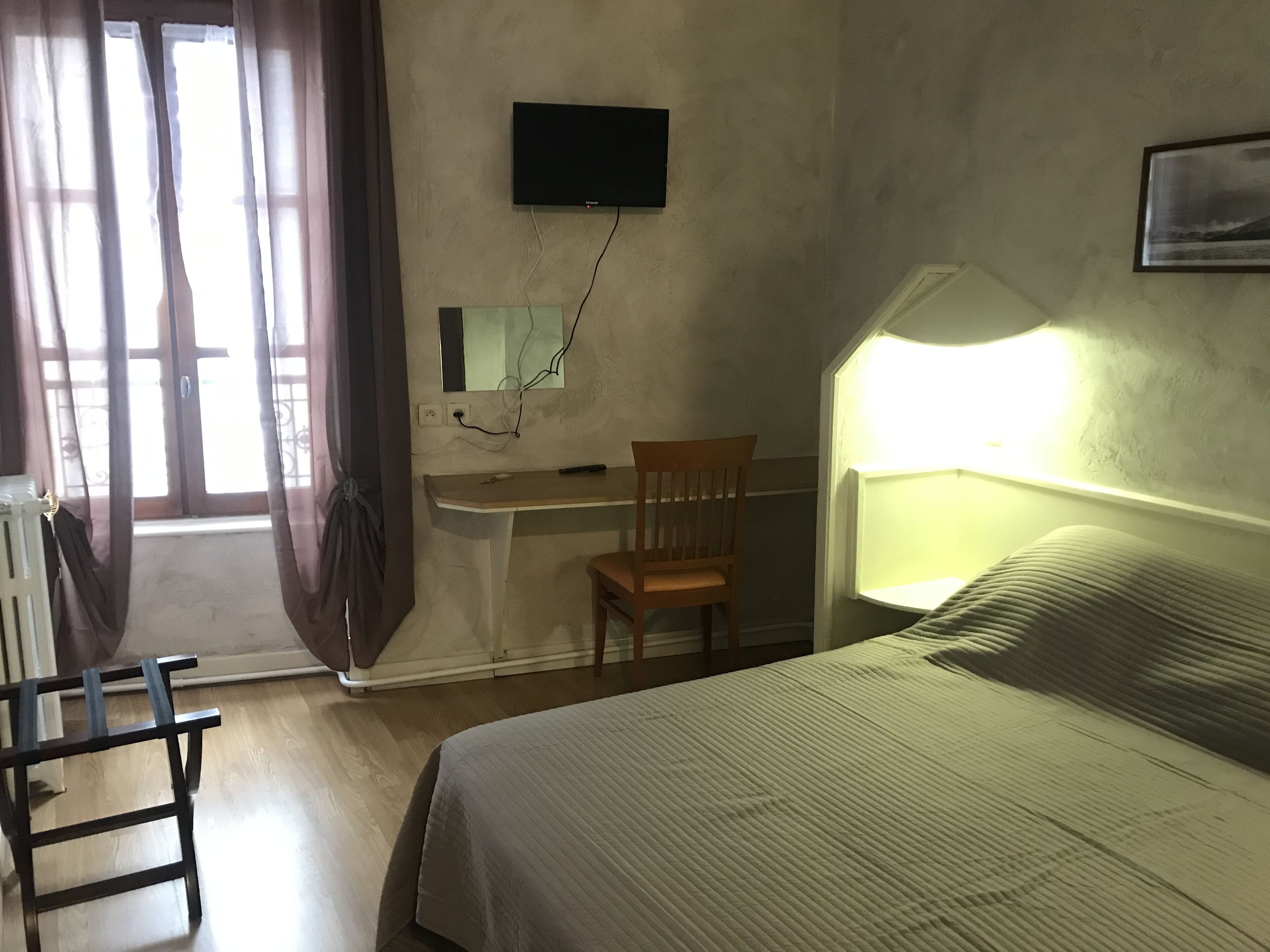 A room with a bed, a desk, a chair, a TV mounted on the wall, and a window with curtains.