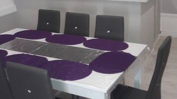 Un coin repas avec une table blanche et des chaises noires, un mur violet et un espace cuisine équipé d’un four à micro-ondes et de placards.