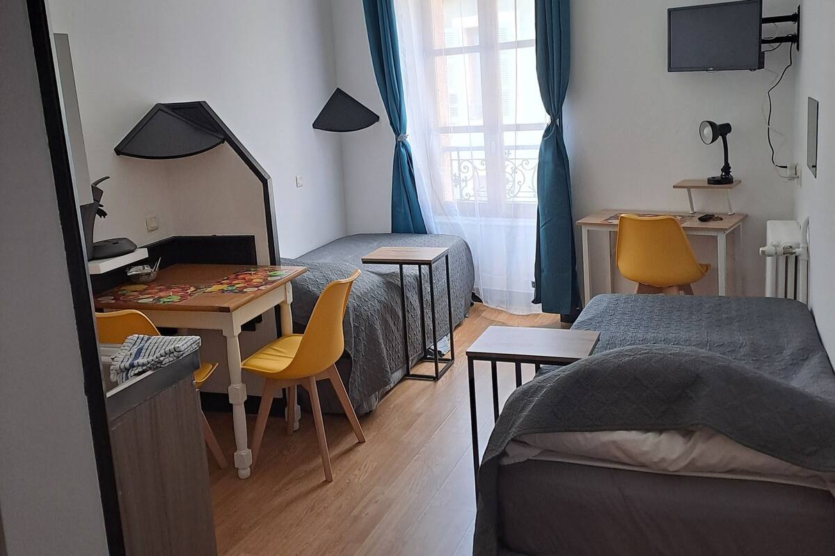 A compact hotel room with a bed, a desk with a chair, a small table, a lamp, and a TV mounted on the wall.