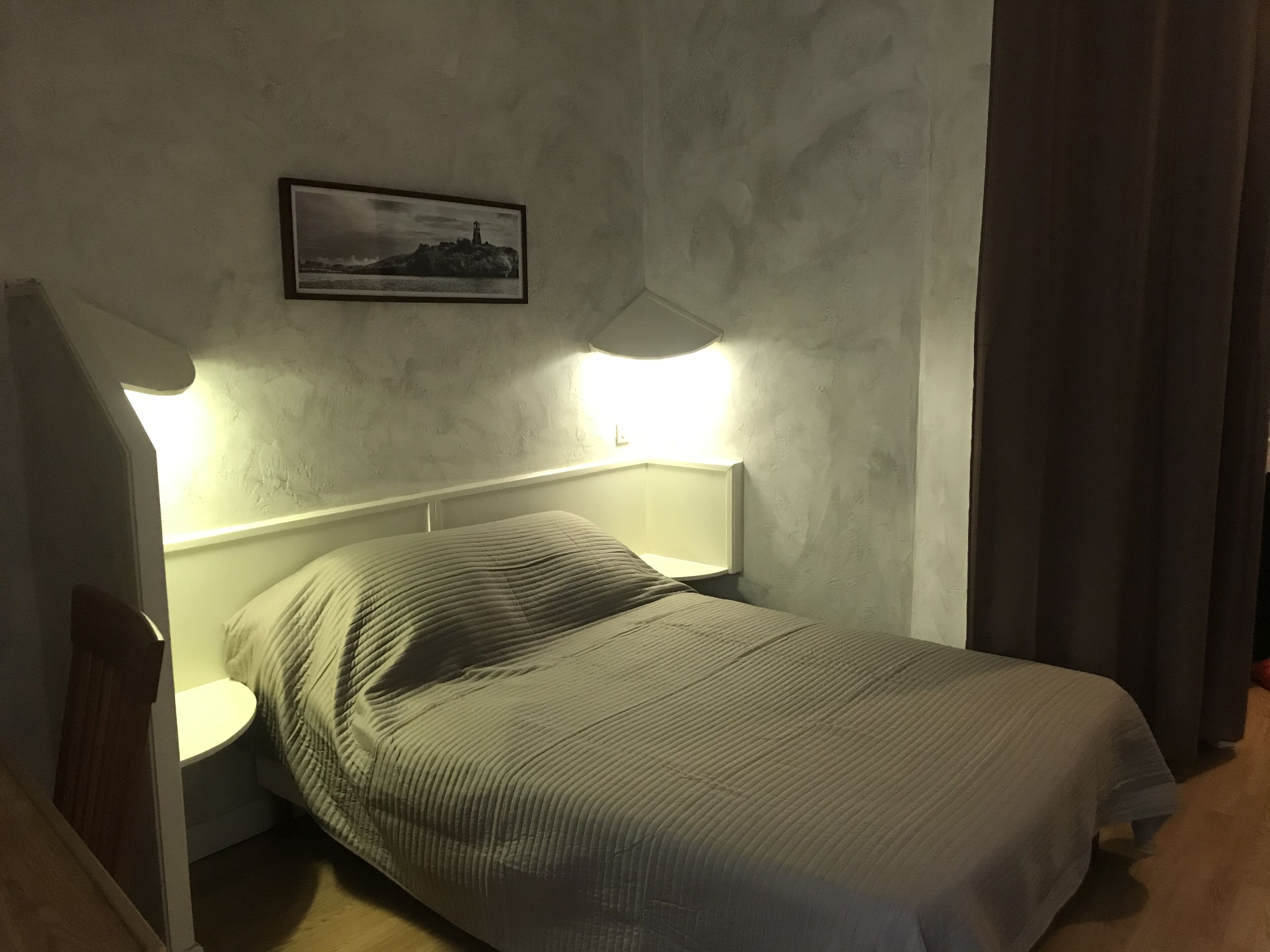 Un lit simple avec une tête de lit, une table de chevet et une lampe fixée au mur.
