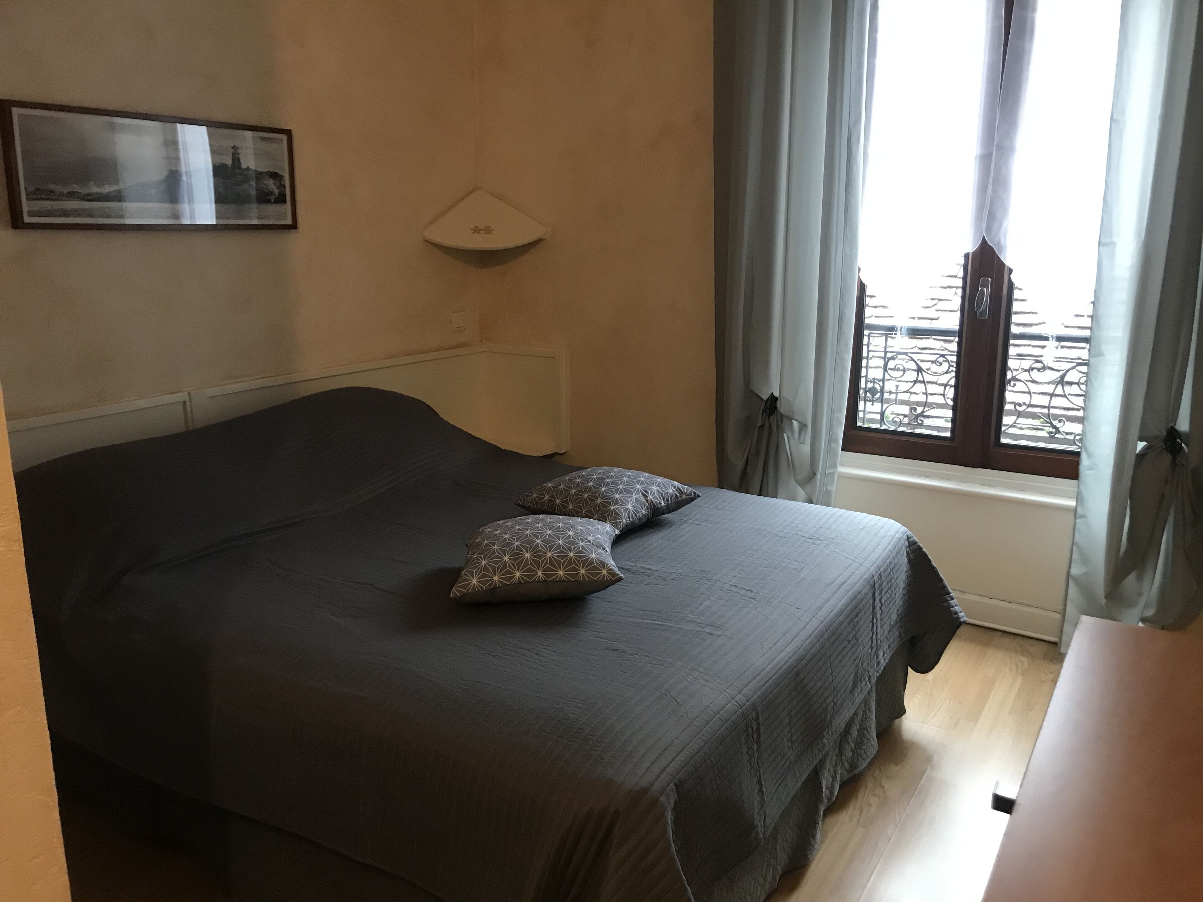 A bedroom with a bed, two pillows, a bedside table, a window with curtains, and a wall-mounted light fixture.