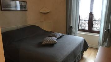 A bedroom with a bed, two pillows, a bedside table, a window with curtains, and a wall-mounted light fixture.
