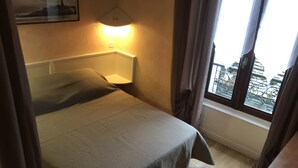 Habitación de hotel con cama, mesita de noche, ventana con cortinas y vistas a edificios.
