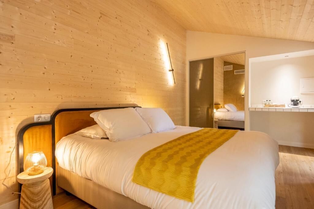 Habitación de hotel con techo de madera, cama con ropa blanca y una manta amarilla, mesita de noche con lámpara, y baño visible a través de una puerta abierta.