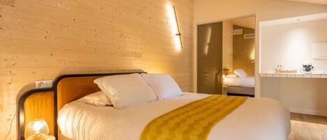 Habitación de hotel con techo de madera, cama con ropa blanca y una manta amarilla, mesita de noche con lámpara, y baño visible a través de una puerta abierta.