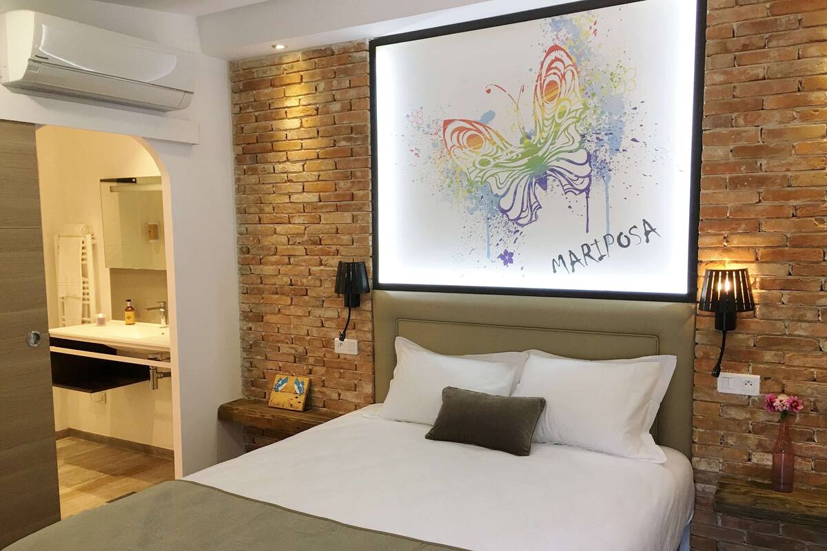 A hotel room with a brick wall, a bed with white linens, a nightstand with a lamp, and a bathroom visible through an archway.