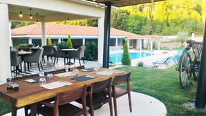 An outdoor patio with a checkered floor, wooden tables, and chairs, overlooking a swimming pool and a forested area.