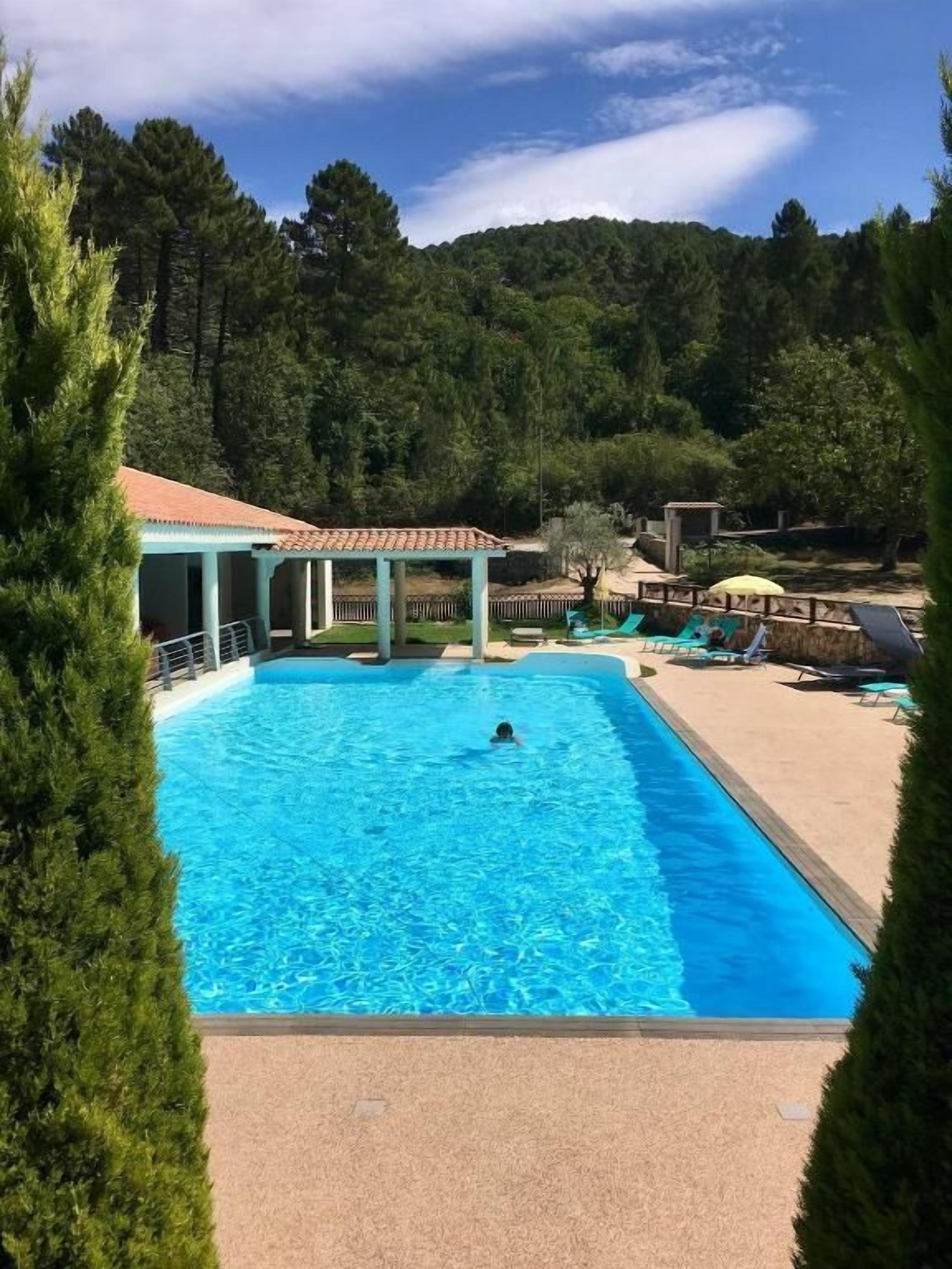 A swimming pool with a person swimming, surrounded by lounge chairs and a covered area.