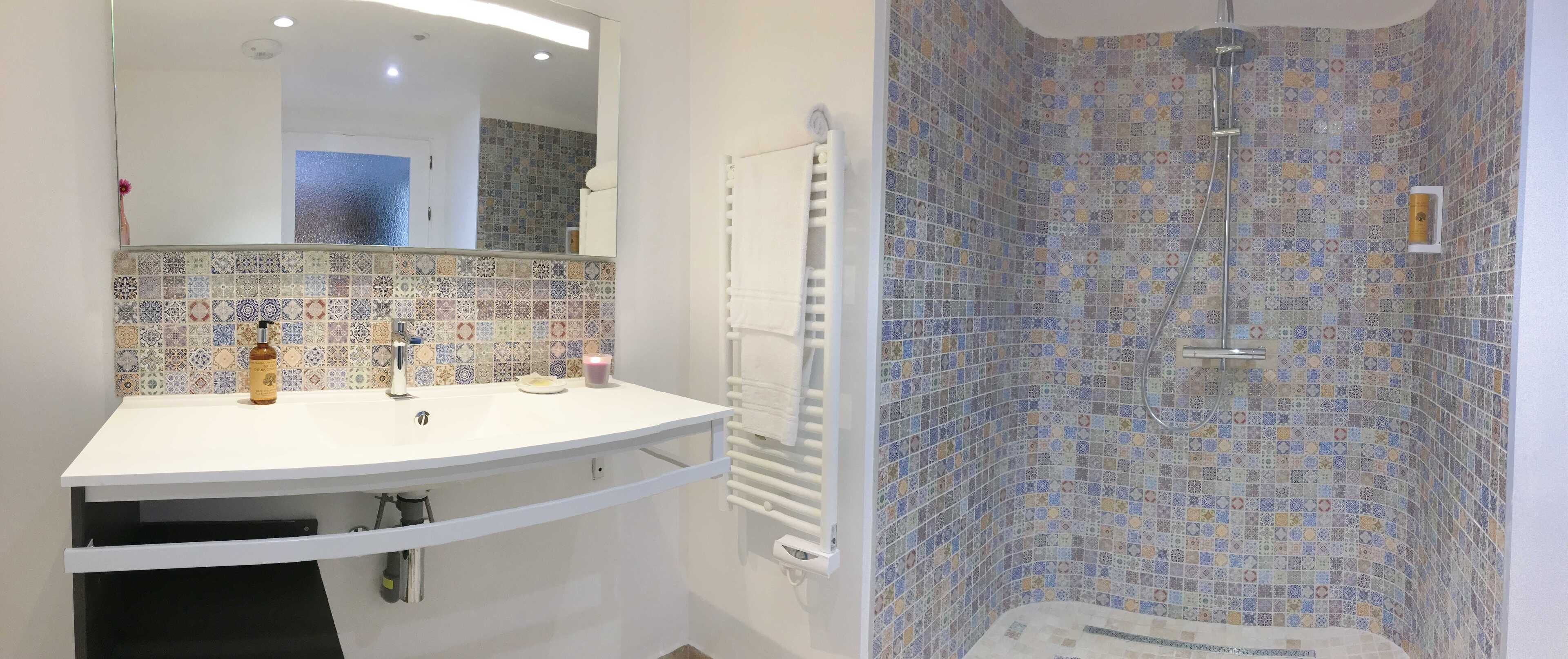 Un baño con lavamanos blanco, espejo, toallero y ducha con azulejos estampados.