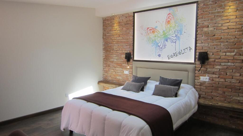 A bedroom with a bed, a brick wall, a colorful artwork, and two wall-mounted lamps.