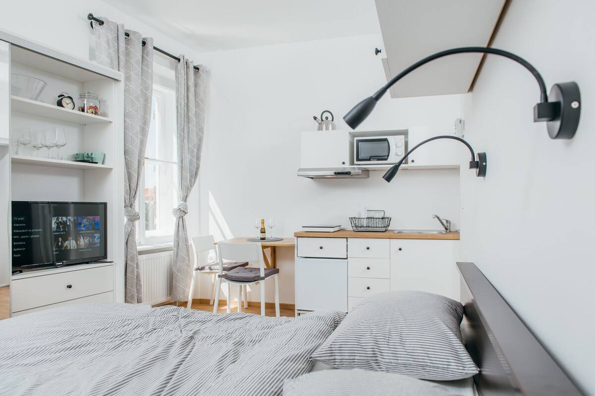 A modern bedroom with a bed, a desk, a chair, a TV, and a shelf with various items.