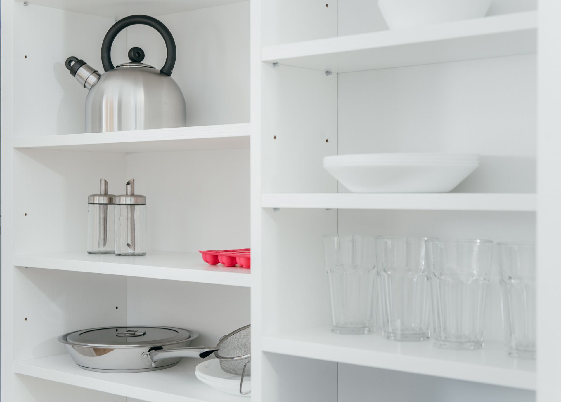 A white kitchen shelf with a kettle, two containers, a pan, a strainer, a red muffin tin, and several clear glasses.