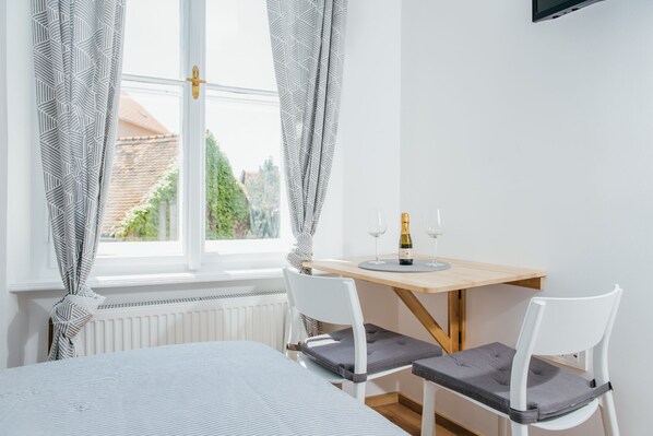A bedroom with a bed, a window with curtains, a table with a bottle and glasses, and two chairs.