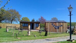 A rustic wooden building with a metal roof, surrounded by a fenced area with a bird feeder and a wooden fence. - Rosie Traditional Gypsy Wagon (Rhayader)