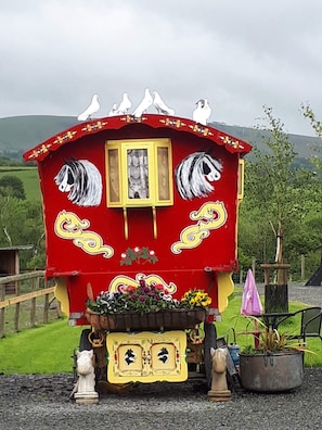 A red carriage with white horses painted on the sides, a small window, and a flower basket at the front. - Rosie Traditional Gypsy Wagon (Rhayader)
