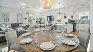 Un salón amplio con una mesa de comedor dispuesta para seis personas, rodeado de muebles modernos y una gran lámpara de araña.
