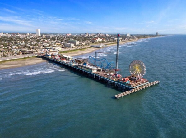 Un muelle se adentra en el agua, con una noria y atracciones, con vistas a una ciudad costera.