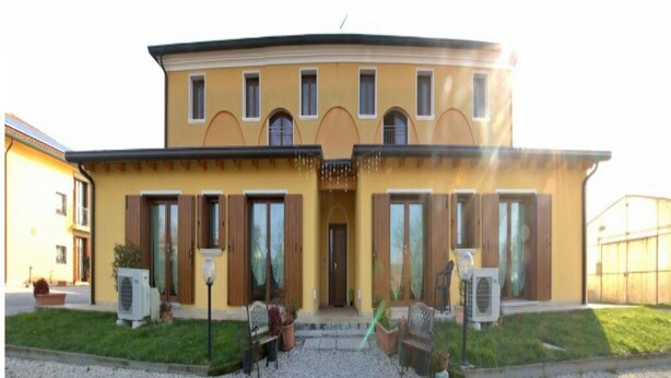 A two-story building with a curved roof, a central entrance with a door, and a small balcony. There are air conditioning units on the ground floor and a bench in front of the building. - Autumn Apartment (Agriturismo Il Melograno) (Venezia)