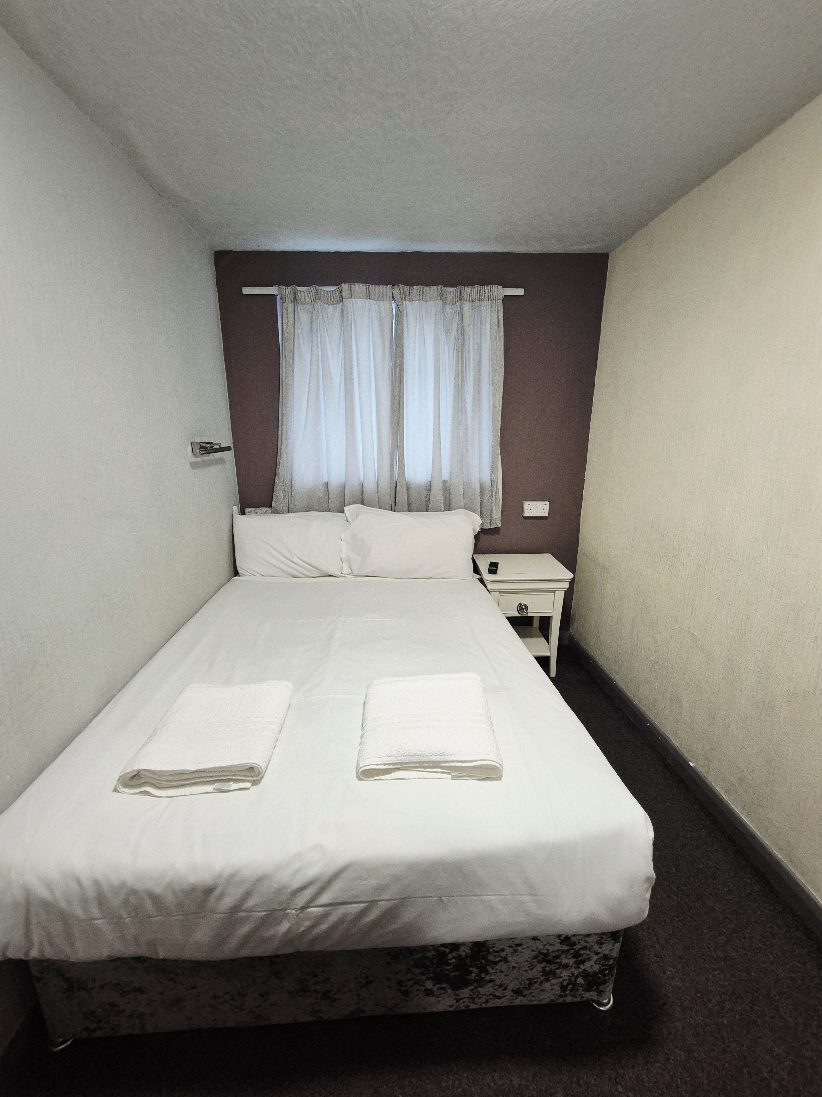 Une chambre d’hôtel avec un lit simple, deux serviettes blanches, une table de chevet, une lampe et une fenêtre avec des rideaux.