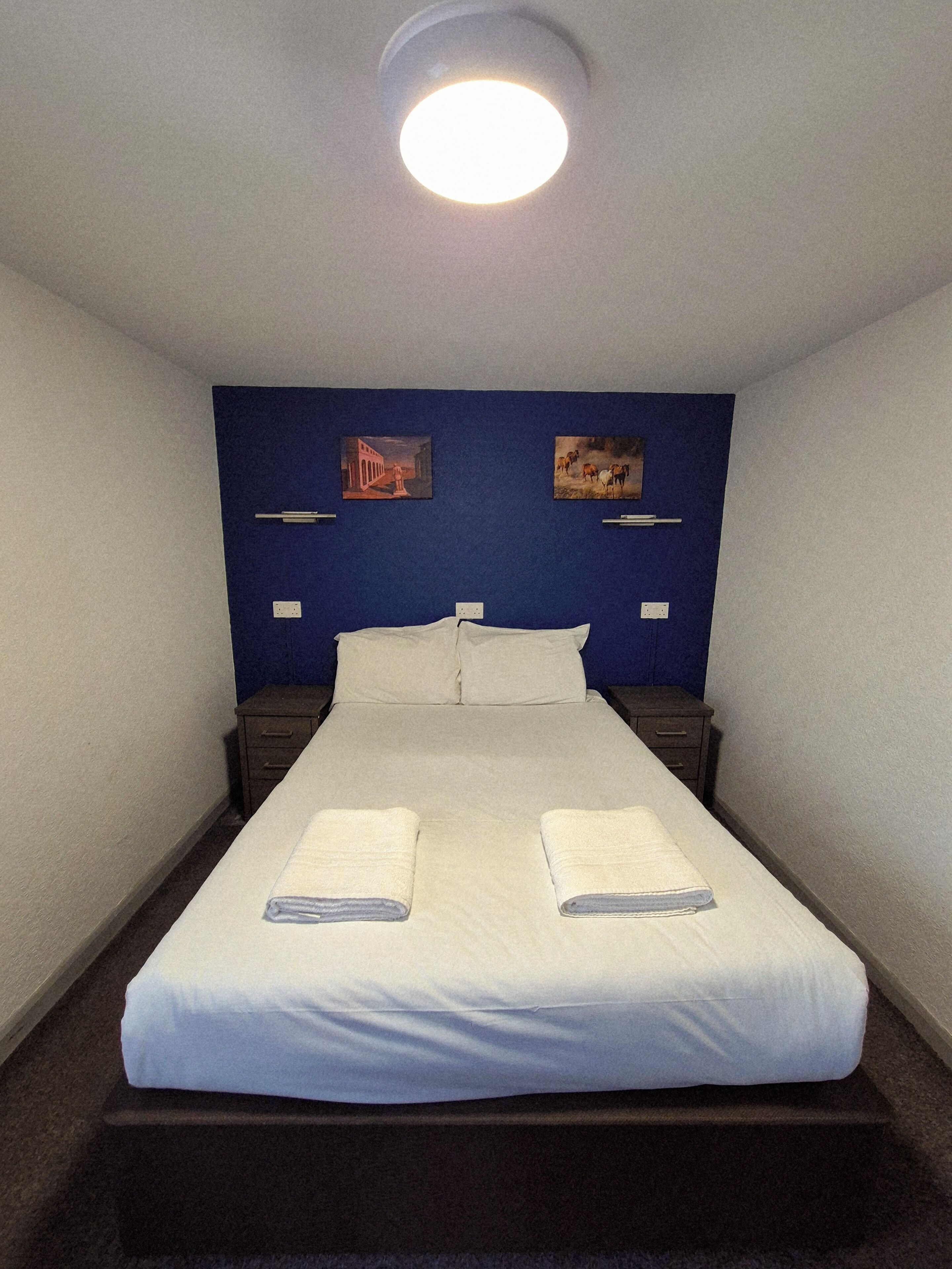Ein Hotelzimmer mit einem großen Bett, zwei Nachttischen, einer Deckenleuchte und zwei an der Wand befestigten Bildern.