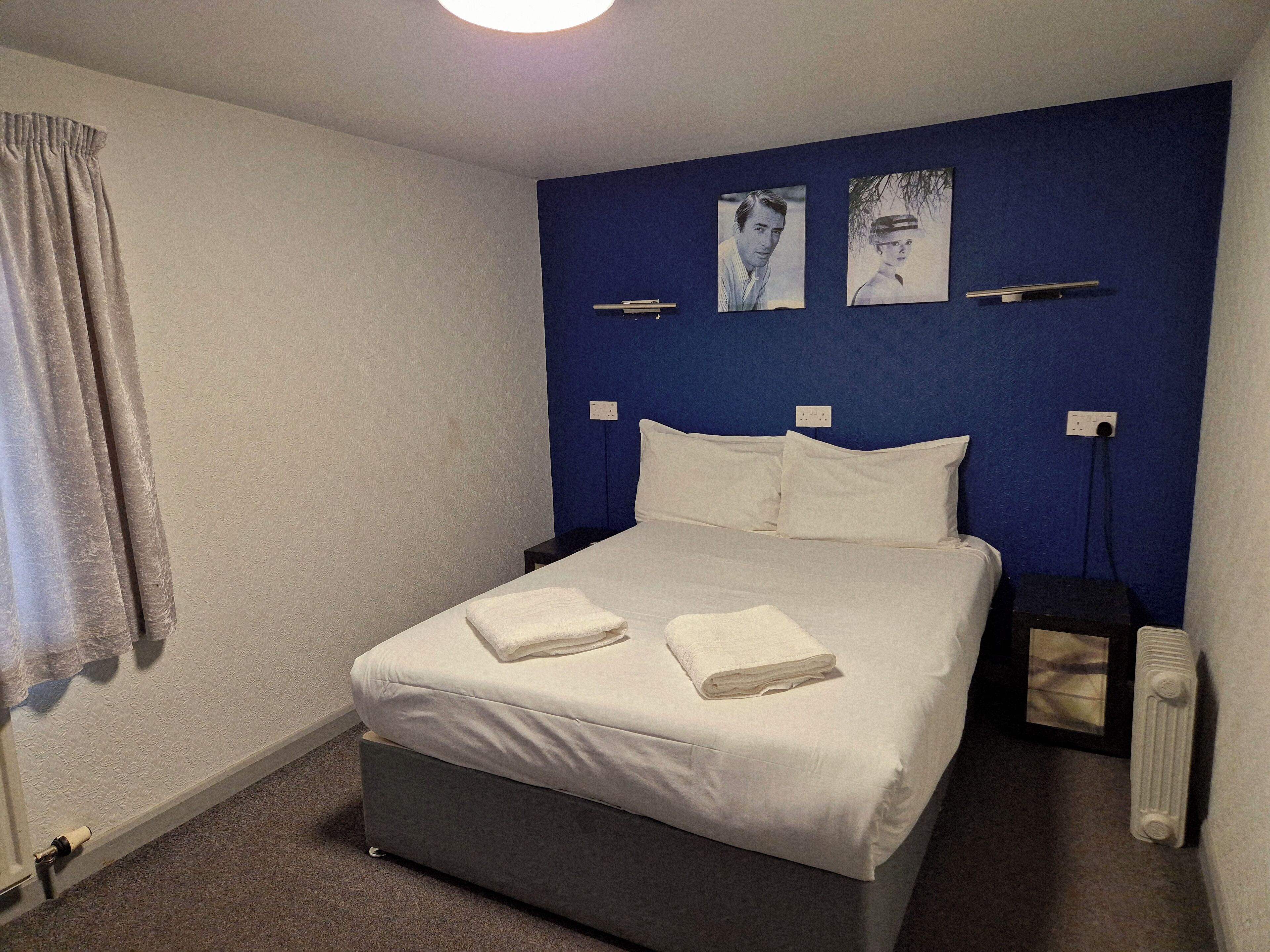 Cama bem arrumada com roupa de cama branca e duas toalhas dobradas, parede de destaque azul e duas mesas de cabeceira com abajures.