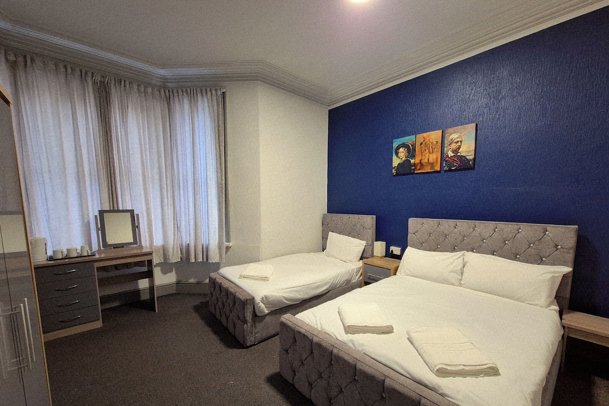 A hotel room with two beds, a desk, a chair, a dresser, and a window with curtains. There are paintings on the wall.