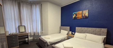 A hotel room with two beds, a desk, a chair, a dresser, and a window with curtains. There are paintings on the wall.