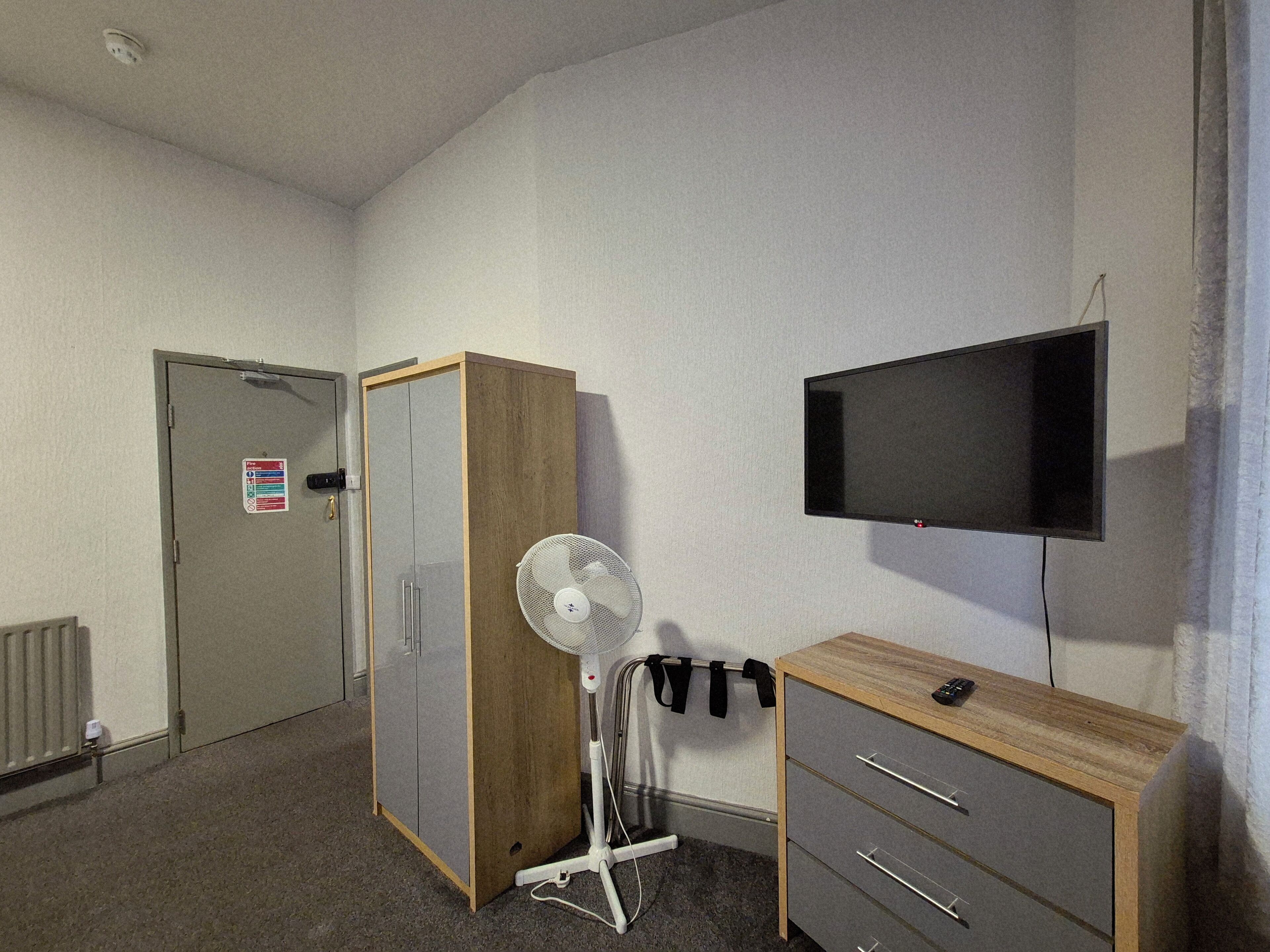 A room with a wardrobe, a desk with drawers, a standing fan, a mounted TV, and a door with a fire safety sign.