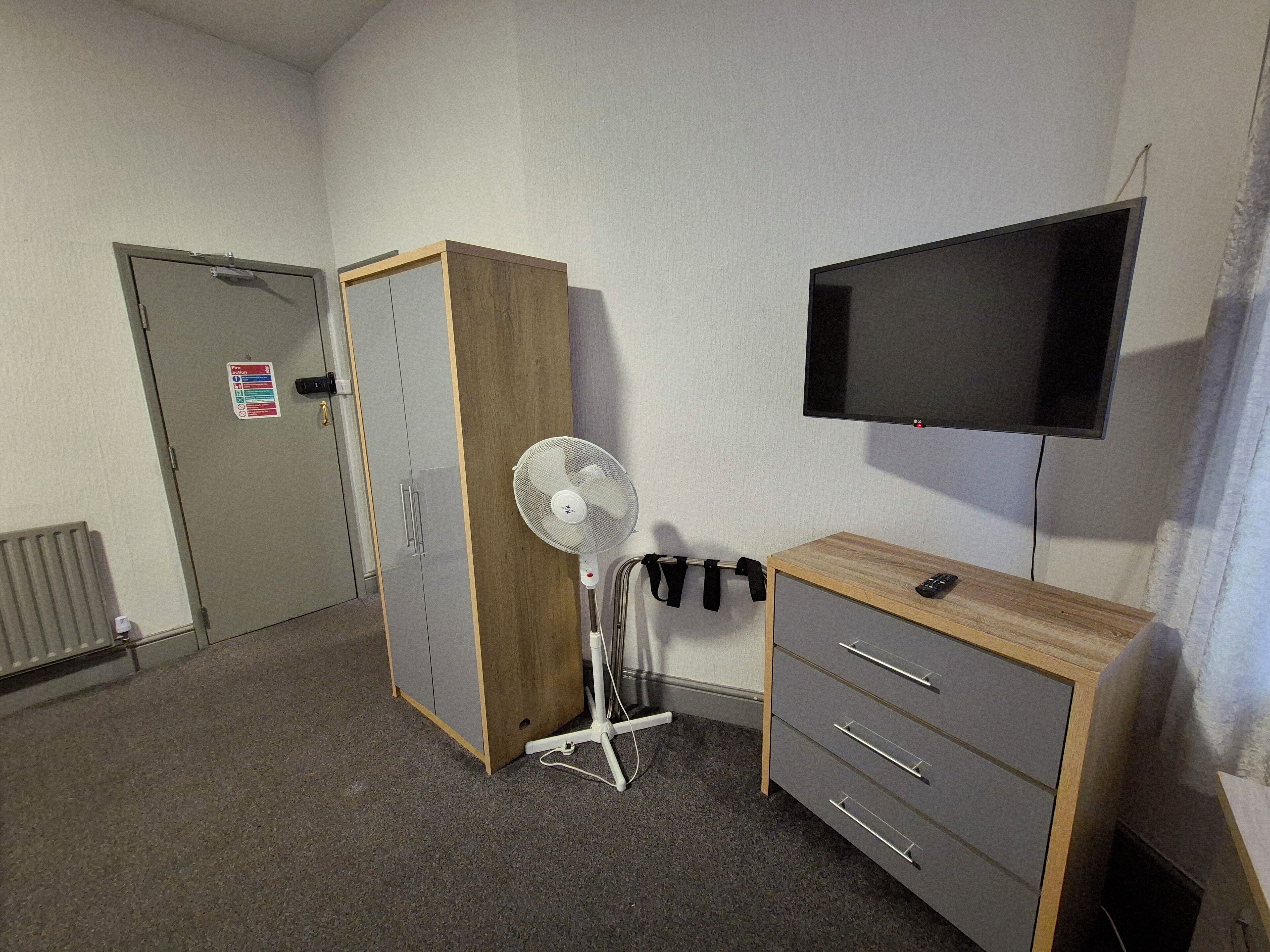 Quarto com guarda-roupa, escrivaninha com gavetas, ventoinha, televisão montada e porta com aviso.